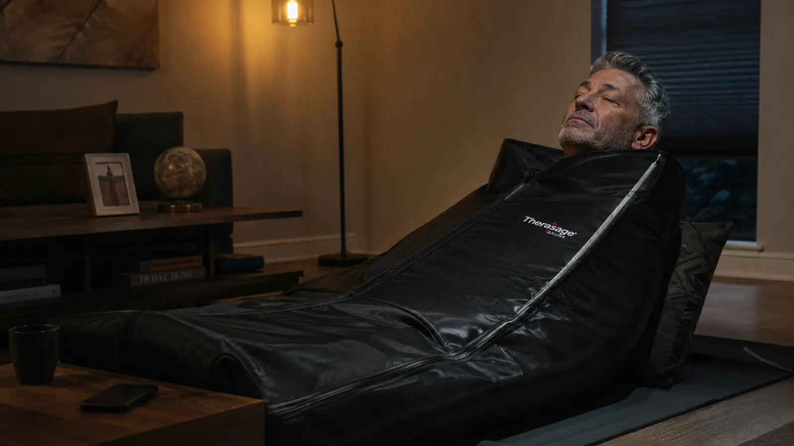 A woman lying on a living room floor mat wrapped in a maroon infrared sauna blanket, only her head and neck visible, soft lamp light in the evening